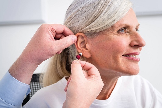 Ältere Frau bekommt ein modernes Hörgerät ins Ohr eingesetzt und lächelt dabei freundlich.